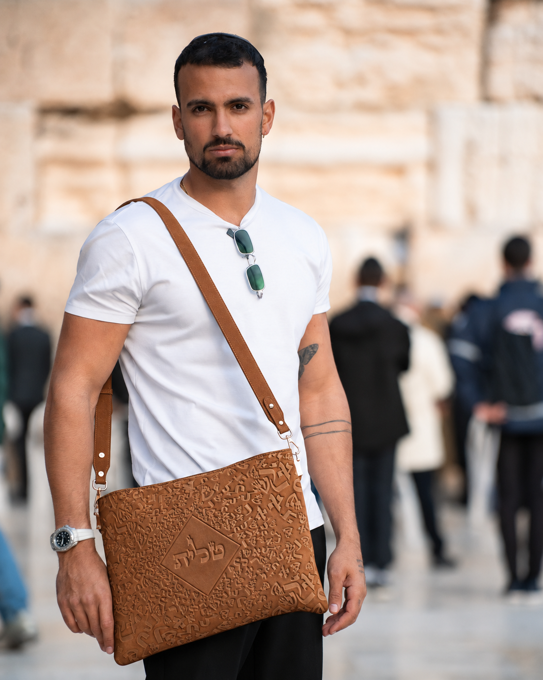 Man in a white shirt wearing the light brown leather tallit bag in front of a stone wall