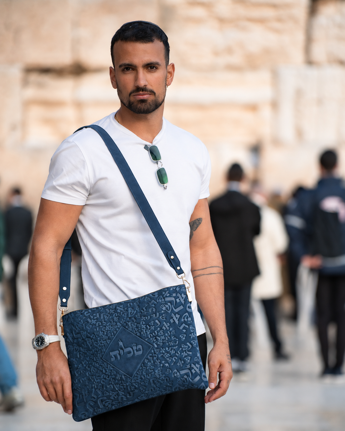 Man in a white shirt wearing the blue leather tallit bag in front of a stone wall